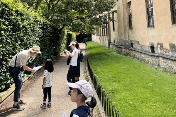 A tour guide explains a map to a young girl whilst her mother takes a photo. They are on a path flanked by a hedge and a stone building.